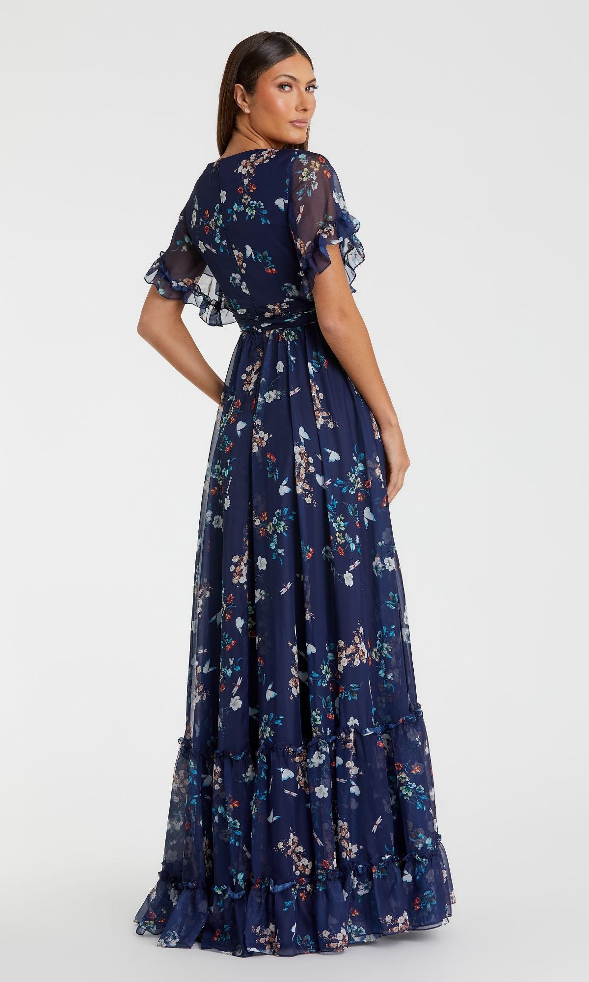 A woman stands facing away from the camera, wearing the Floral-Print Long Formal Dress: 70241 Mac Duggal—a dark blue gown with floral print, short ruffled sleeves, and a tiered hem.